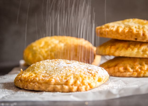 sprinkling powdered sugar on a hand pie with 3 stacked and 2 leaning on each other in the back ground