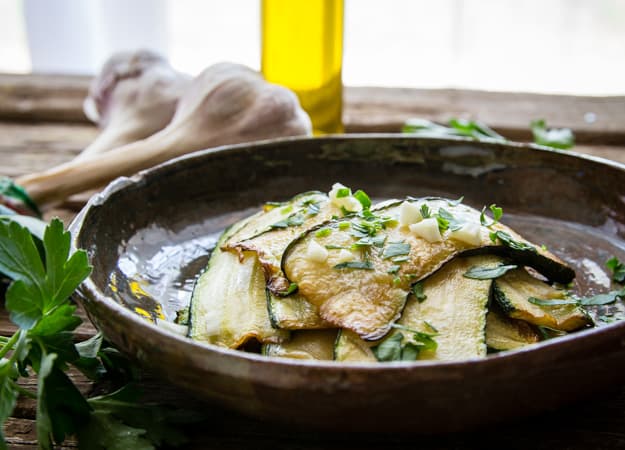 Italian marinated zucchini in a bowl with chopped parsley and garlic, a bottle of oil in the background