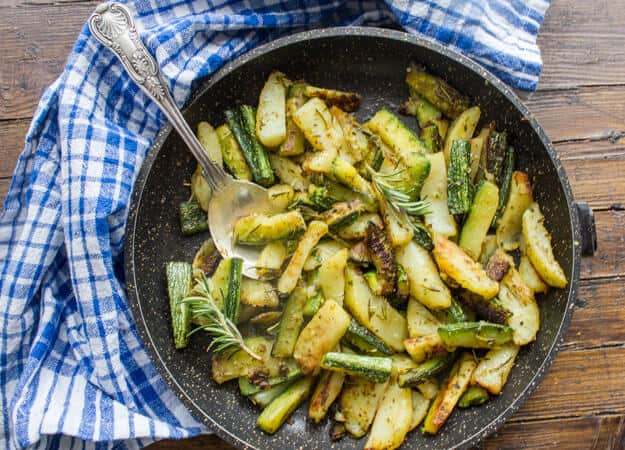 Geröstete Zucchini-Kartoffeln in einer schwarzen Pfanne mit einem silbernen Servierlöffel und einer blau-weißen Serviette