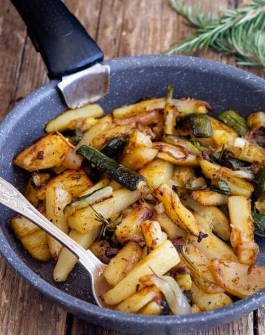 Zucchini and potatoes in a black pan with a silver spoon.