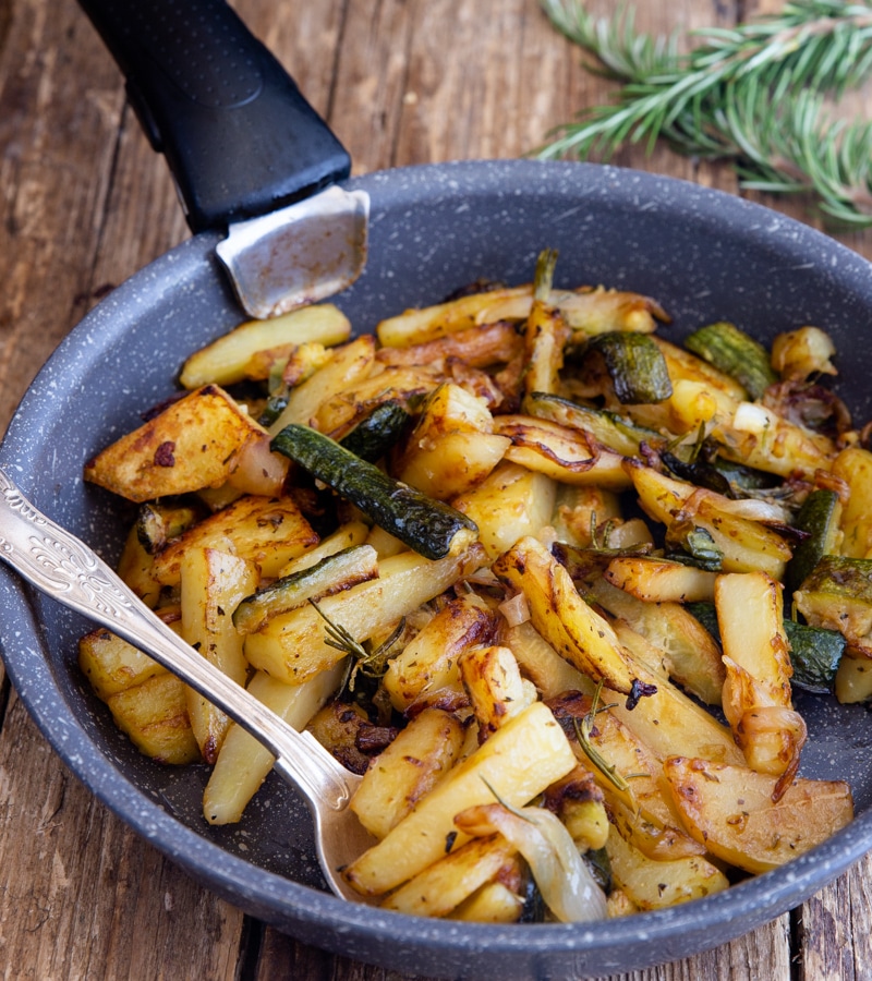 Zucchini and potatoes in a black pan with a silver spoon.