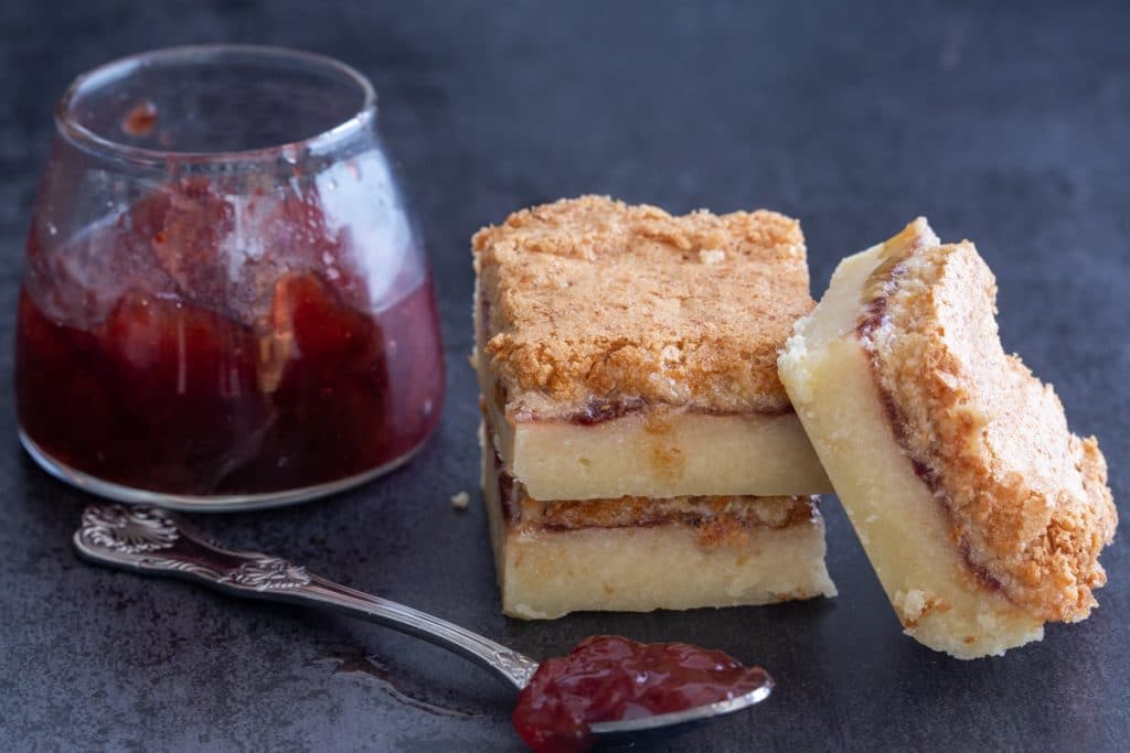 2 Coconut jam squares stacked with one leaning and jam in glass jar with a silver spoon.
