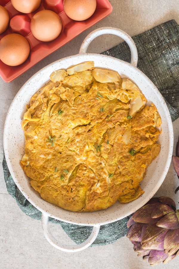 frittata in a white baking dish with eggs in a dish and an artichoke