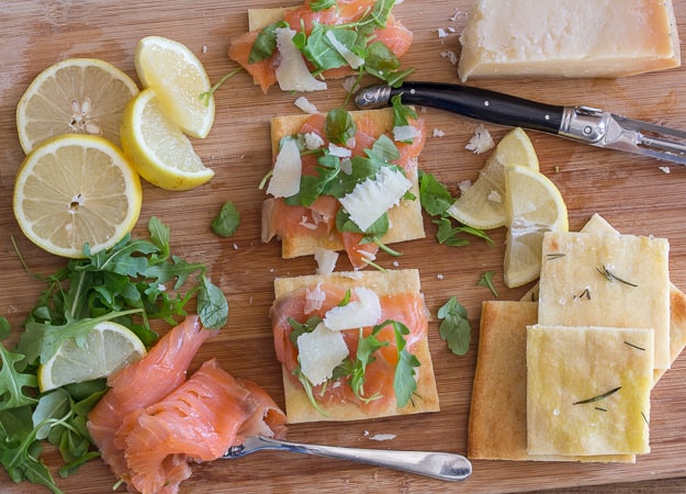 smoked salmon slices, parmesan cheese chunks, rucola leaves, lemon slices and a knife on a wooden board