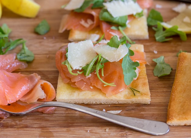 smoked salmon slices on squares of pizza with flakes of parmesan and lemon slices on a board