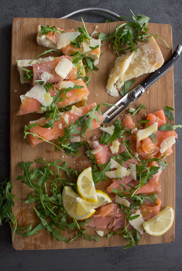 smoked salmon slices, parmesan cheese chunks, rucola leaves, lemon slices and a knife on a wooden board