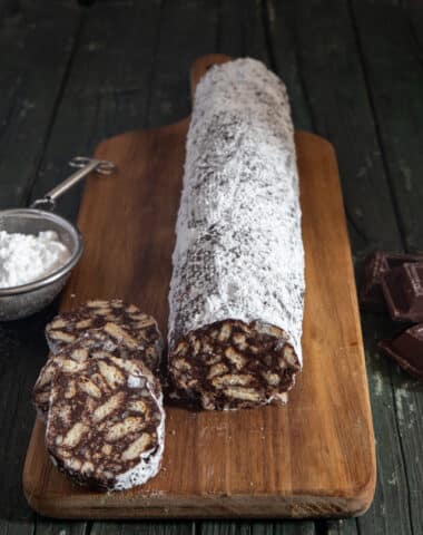 The salami and 3 pieces cut on a wooden board with powdered sugar in a sieve and 3 chunks of chocolate.
