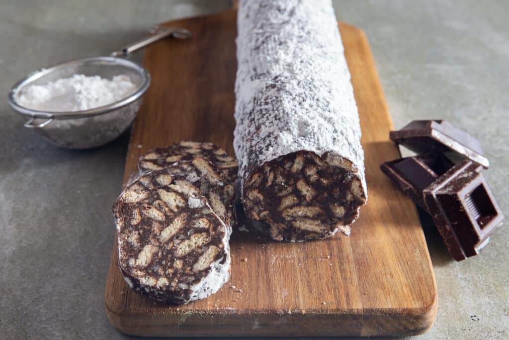 The salami and 3 pieces cut on a wooden board with powdered sugar in a sieve and 3 chunks of chocolate.