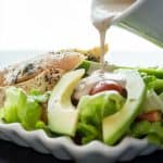 pouring creamy italian dressing on grilled chicken with salad on a white plate