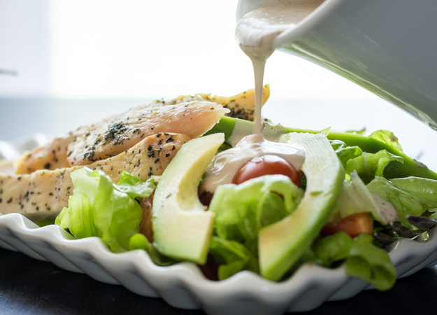 pouring creamy italian dressing on grilled chicken with salad on a white plate 