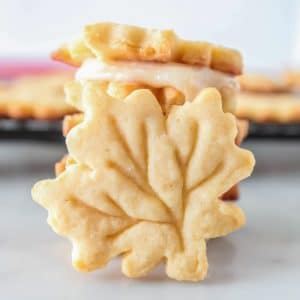 maple leaf cookies stacked with one leaning against them and some on a wire rack with one leaning in the background