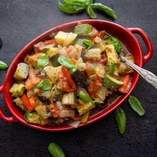 top view of cooked ratatouille in a red casserole dish on a black board with basil leaves