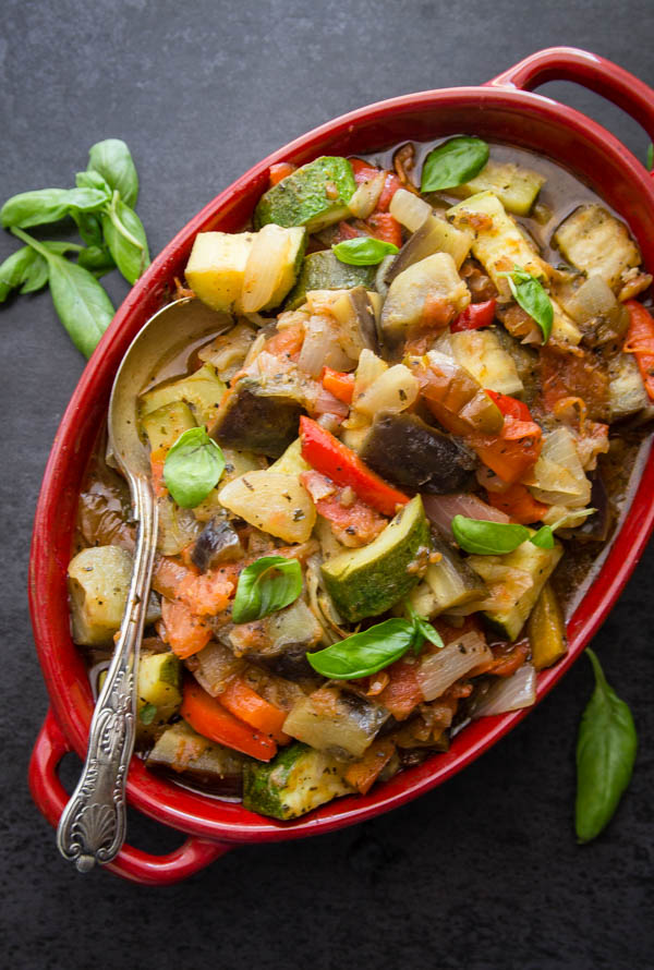 cooked ratatouille in a red oblong casserole serving dish with a silver spoon and basil leaves