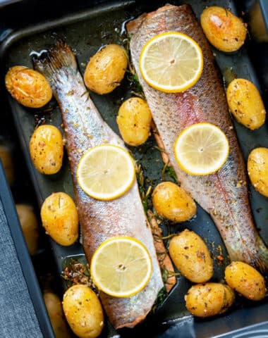 Two baked trouts with potatoes in a black pan.