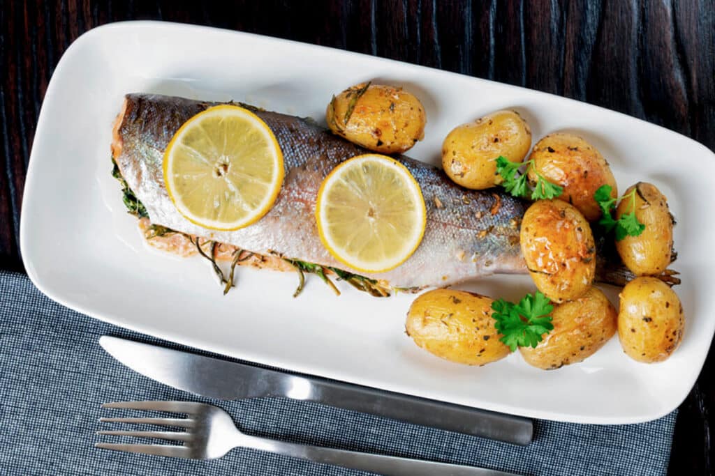 Trout on a white dish with lemon slices and potatoes.