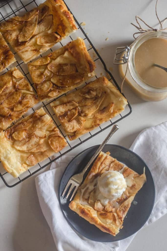 Cinnamon apple tart on a wire rack with a slice on a black plate.