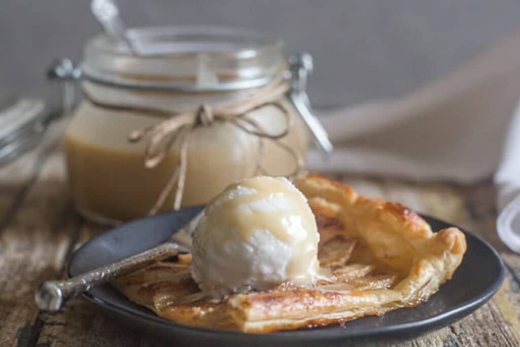 A slice of apple tart on a black plate with a scoop of ice cream.