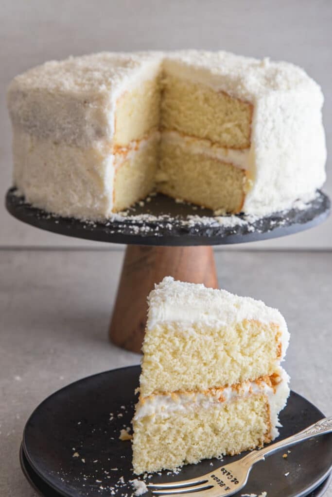 Coconut cake on a cake stand with a slice on a black plate.