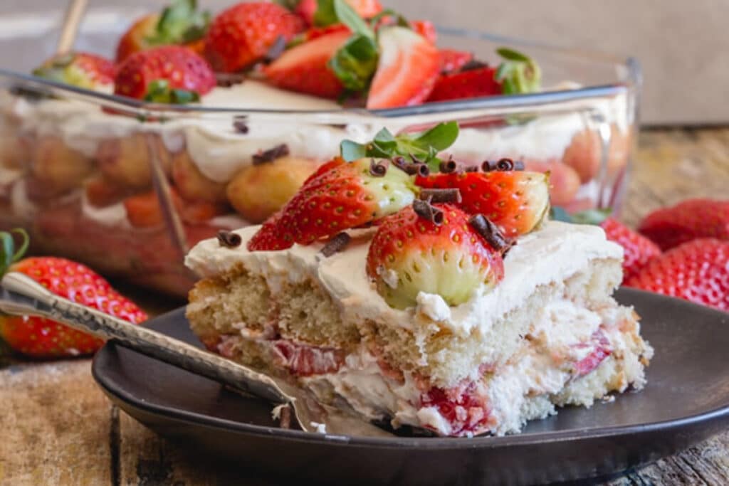 Strawberry tiramisu in a glass pan with a slice on a black plate.