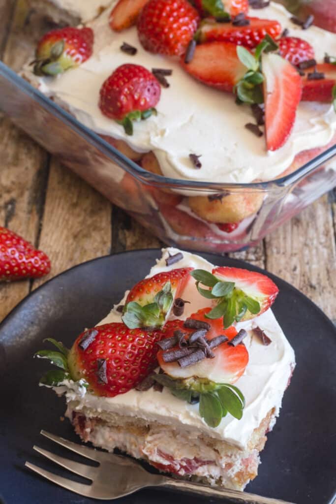 Strawberry tiramisu in a glass pan with a slice on a black plate.