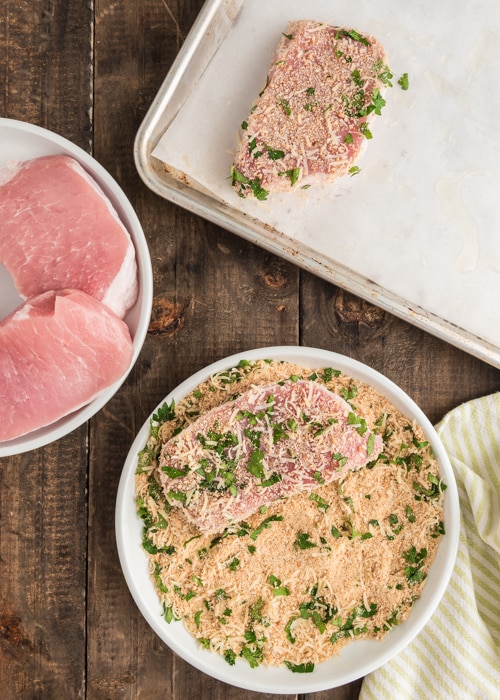 Pork chops on a white plate, crumbs in a bowl and a pork chop on the prepared baking sheet.