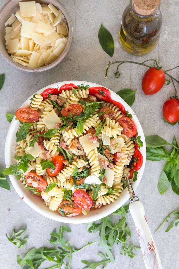 pasta salad on a white board with parmesan flakes in a bowl and olive oil and tomatoes