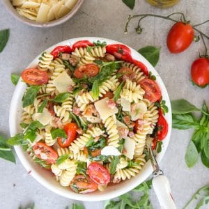 pasta salad on a white board with parmesan flakes in a bowl and olive oil and tomatoes