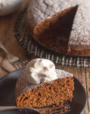 Gingerbread cake on a black wire plate with a slice on a black plate & a dollop of whipped cream.