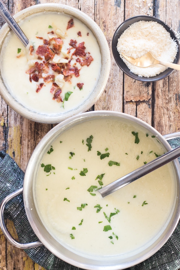 cauliflower soup in a silver pot with a ladle and in a bowl with chopped bacon and parmesan cheese in a small black bowl
