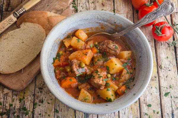 beef stew in a blue bowl with slices of bread and fresh tomatoes in the background