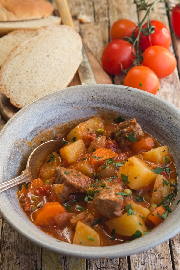beef stew in a blue bowl with slices of bread and fresh tomatoes in the background