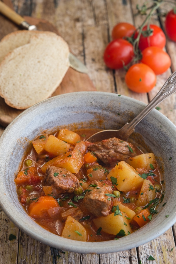 beef stew in a blue bowl with slices of bread and fresh tomatoes in the background