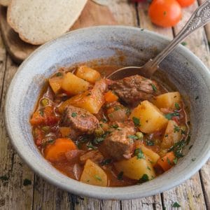 beef stew in a blue bowl with slices of bread and fresh tomatoes in the background