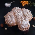 Colomba on a black board with 2 orange slices, almonds and powdered sugar in a sieve.