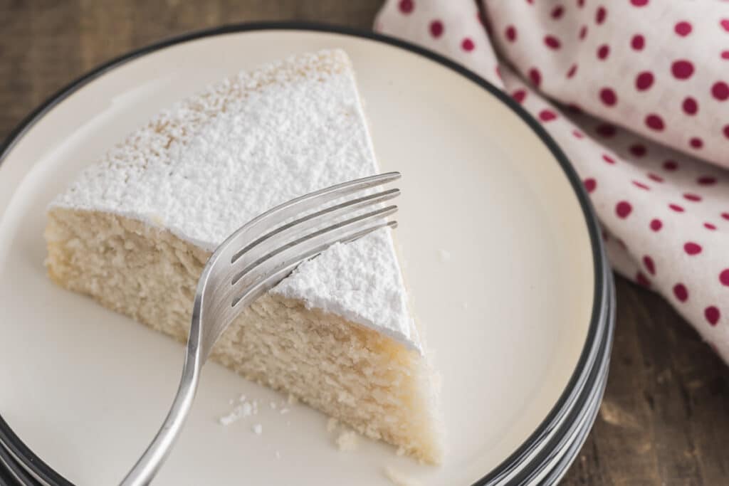 A slice of cake on a white plate cutting a bite with a fork.