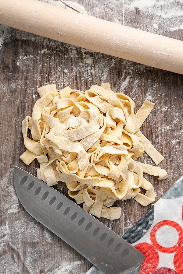 homemade pasta noodles on a brown board with a rolling pin and the blade of a knife