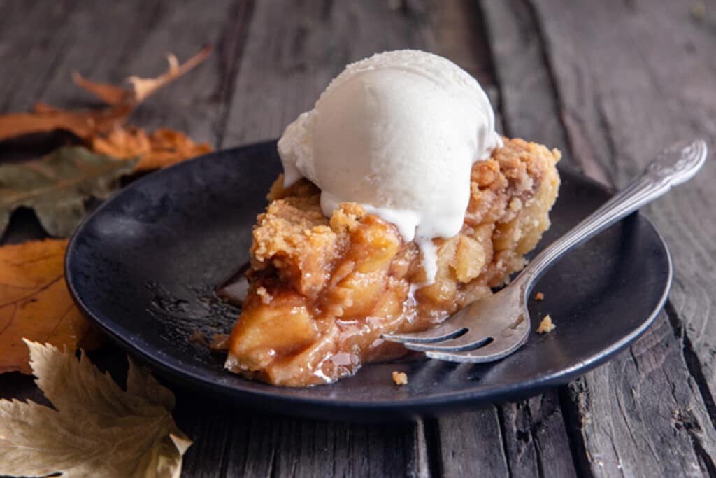 A slice of apple pie on a black plate with a scoop of ice cream.