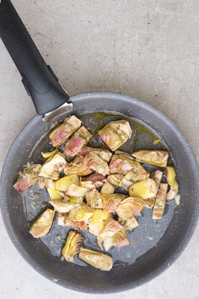 Raw artichokes, oil, garlic and spices in a black pan.