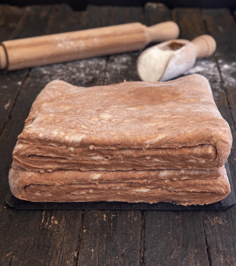 puff pastry with a rolling pin and small scoop of flour on a brown board.