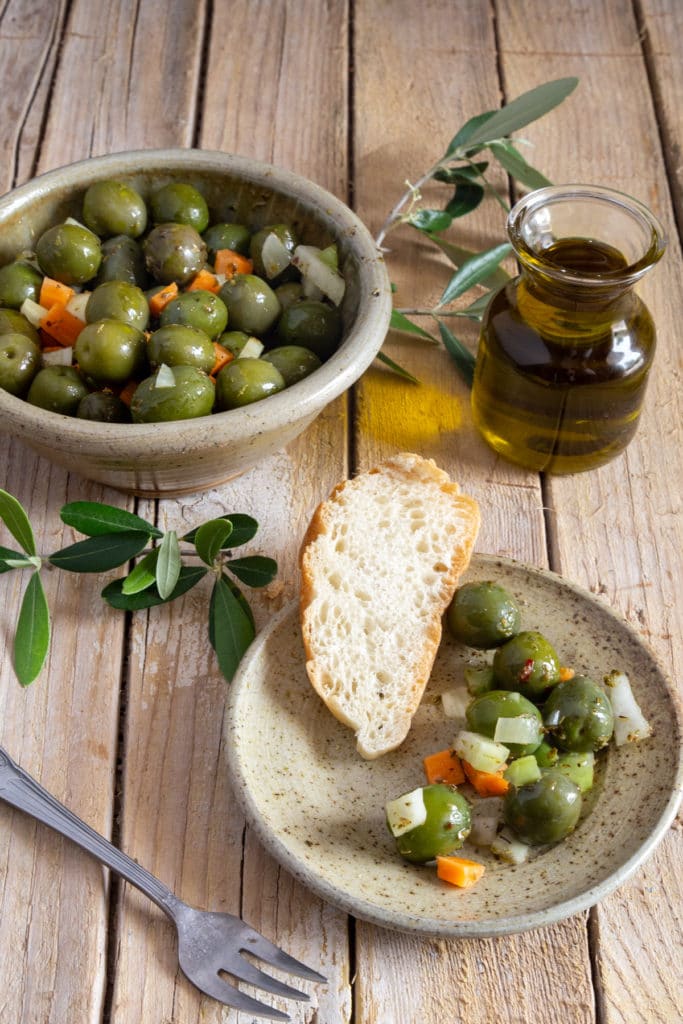 Olives in a grey bowl with some on a grey plate with a slice of bread.