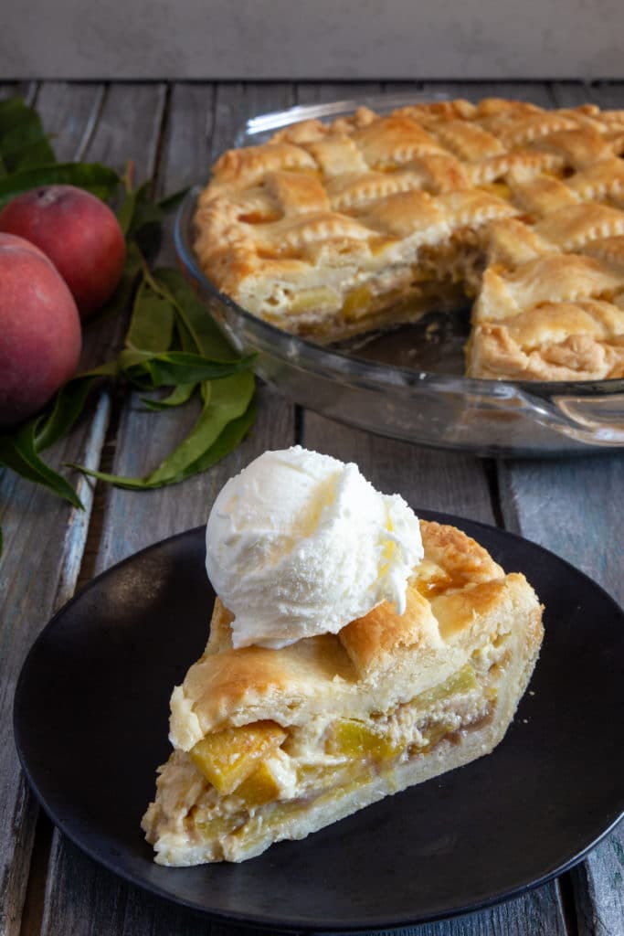 peach pie in a glass dish with a slice & a scoop of ice cream on a black plate.