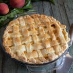 Peach pie in a glass plate, with 2 peaches and leaves on a wooden board.
