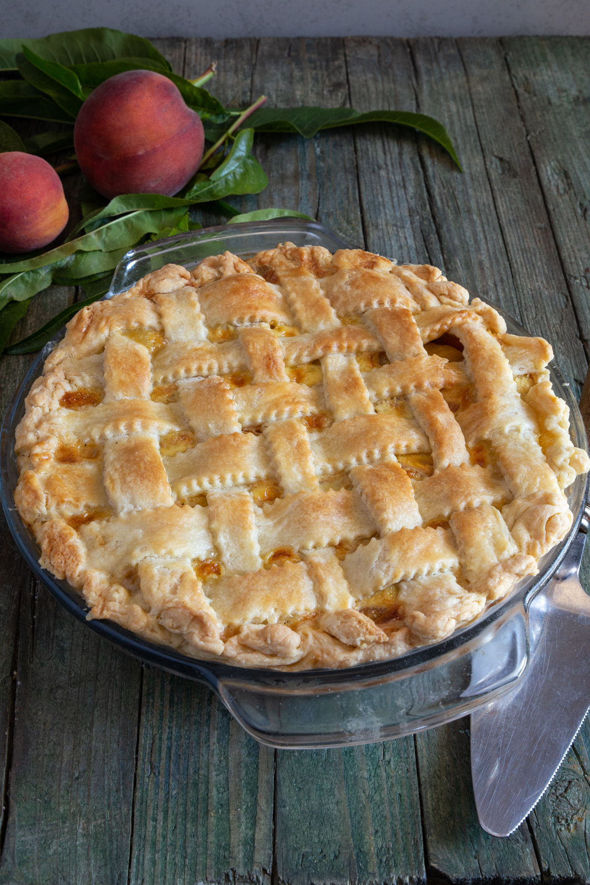 Peach pie in a glass plate, with 2 peaches and leaves on a wooden board.