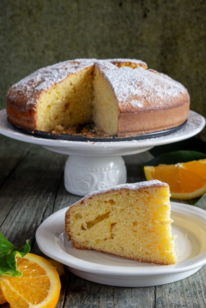 Orange cake on a white cake stand with a slice cut on a white plate.