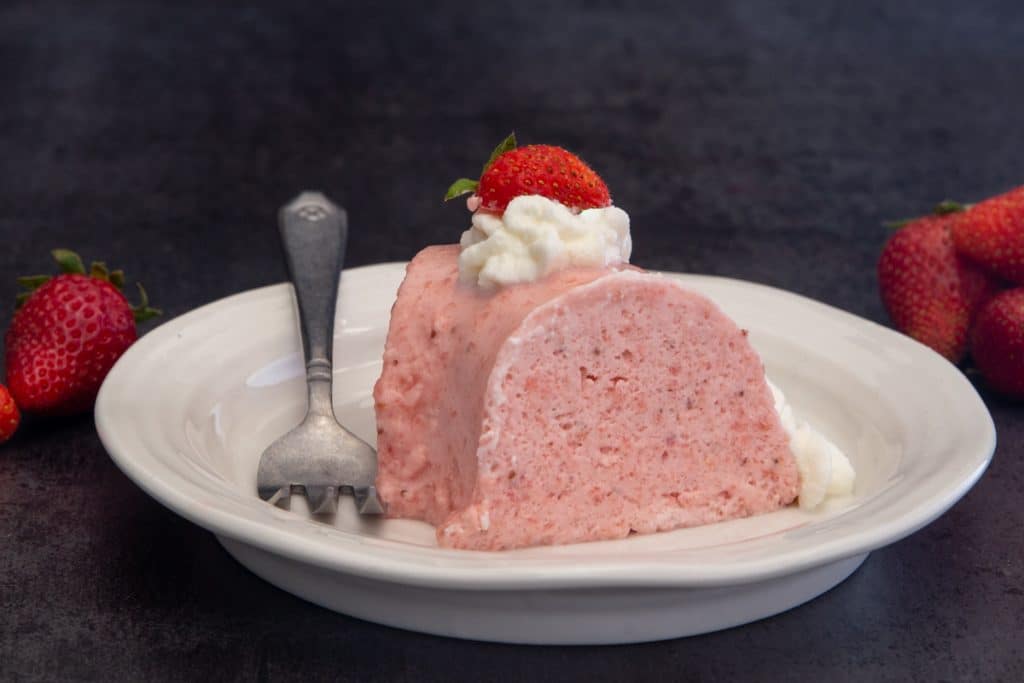 A slice of Italian bavarese on a white plate with a dollop of whipped cream and a strawberry on top.