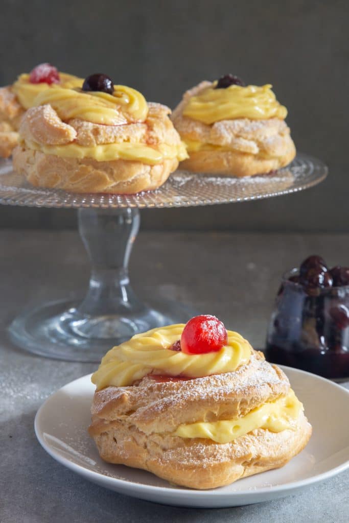 Zeppole on a glass cake stand and one on a white plate.