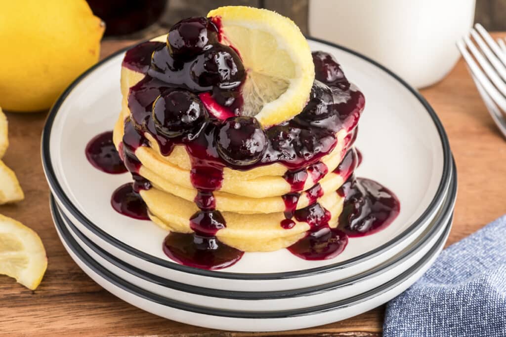 Stacked pancakes on a plate with blueberry sauce and sliced lemons.