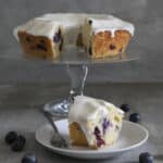 Cake on a glass cake stand with a slice on a white plate.