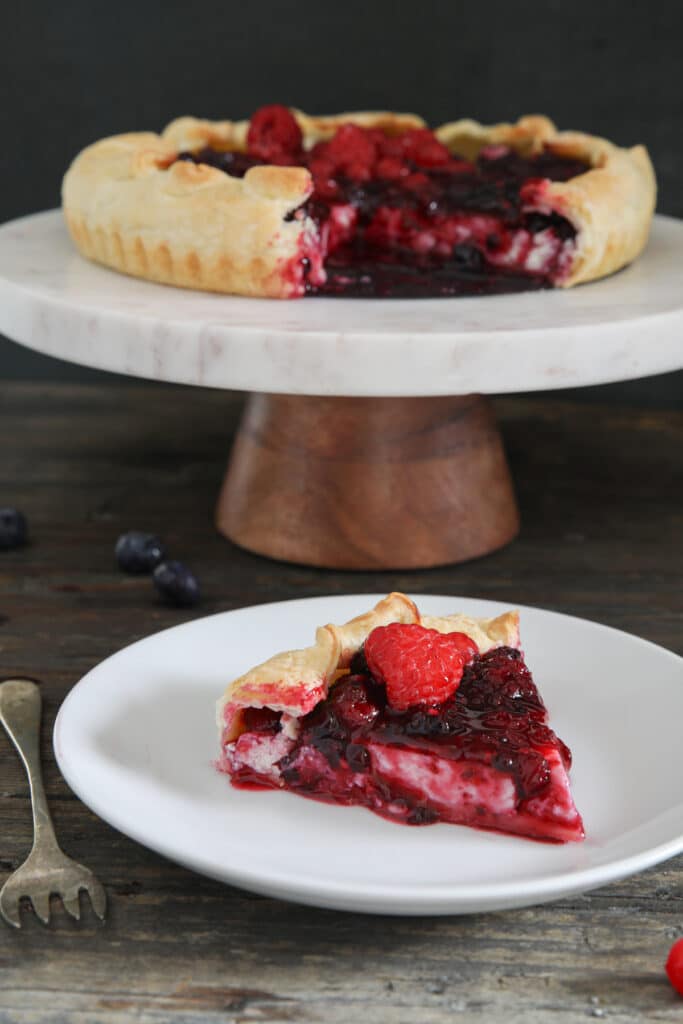 The berry ricotta pie on a cake stand and a slice on a white plate.