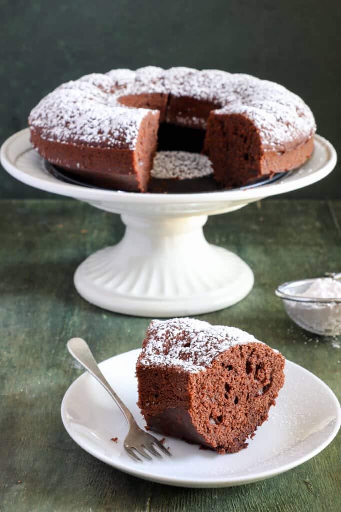 Cake on a white cake stand with a slice on a white plate.